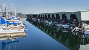 Un puerto deportivo con barcos amarrados en aguas tranquilas, una estructura cubierta a la derecha y un cielo despejado al fondo.