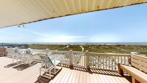 A wooden deck with a white railing, outdoor furniture, and a view of the beach and ocean.