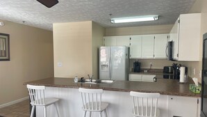 Una cocina con encimera de granito, taburetes de bar blancos, un refrigerador, un microondas y una cafetera.