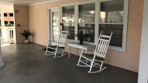 Un porche con dos mecedoras y una mesa, paredes color beige y ventanas grandes con persianas.
