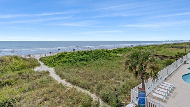 A beach view with a pathway leading to a pool area, surrounded by greenery and a wooden fence.
