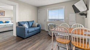A compact living space with a blue sofa, a dining table with white chairs, a ceiling fan, and a window with blinds.