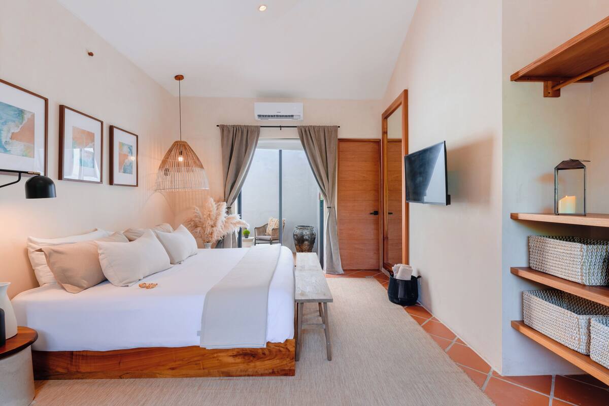 A modern bedroom with a large bed, a bedside table, a desk, a chair, a TV, and a shelf with baskets.