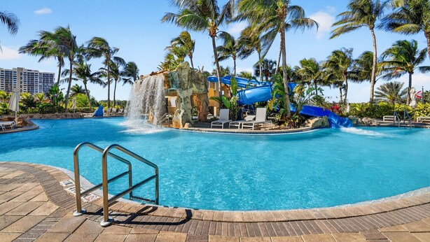 A swimming pool with a water slide, palm trees, and a waterfall feature.