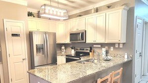 A kitchen with white cabinets, a stainless steel refrigerator, a microwave, a coffee maker, and a granite countertop. - Balcony to beach in minutes! Orange Beach tranquility all year at great rates! (Orange Beach)