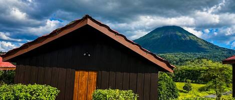A wooden cabin with a sloped roof, surrounded by greenery and a mountain in the background.