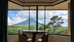 A room with a bed, a table, and chairs, large windows with a view of a mountain and greenery.