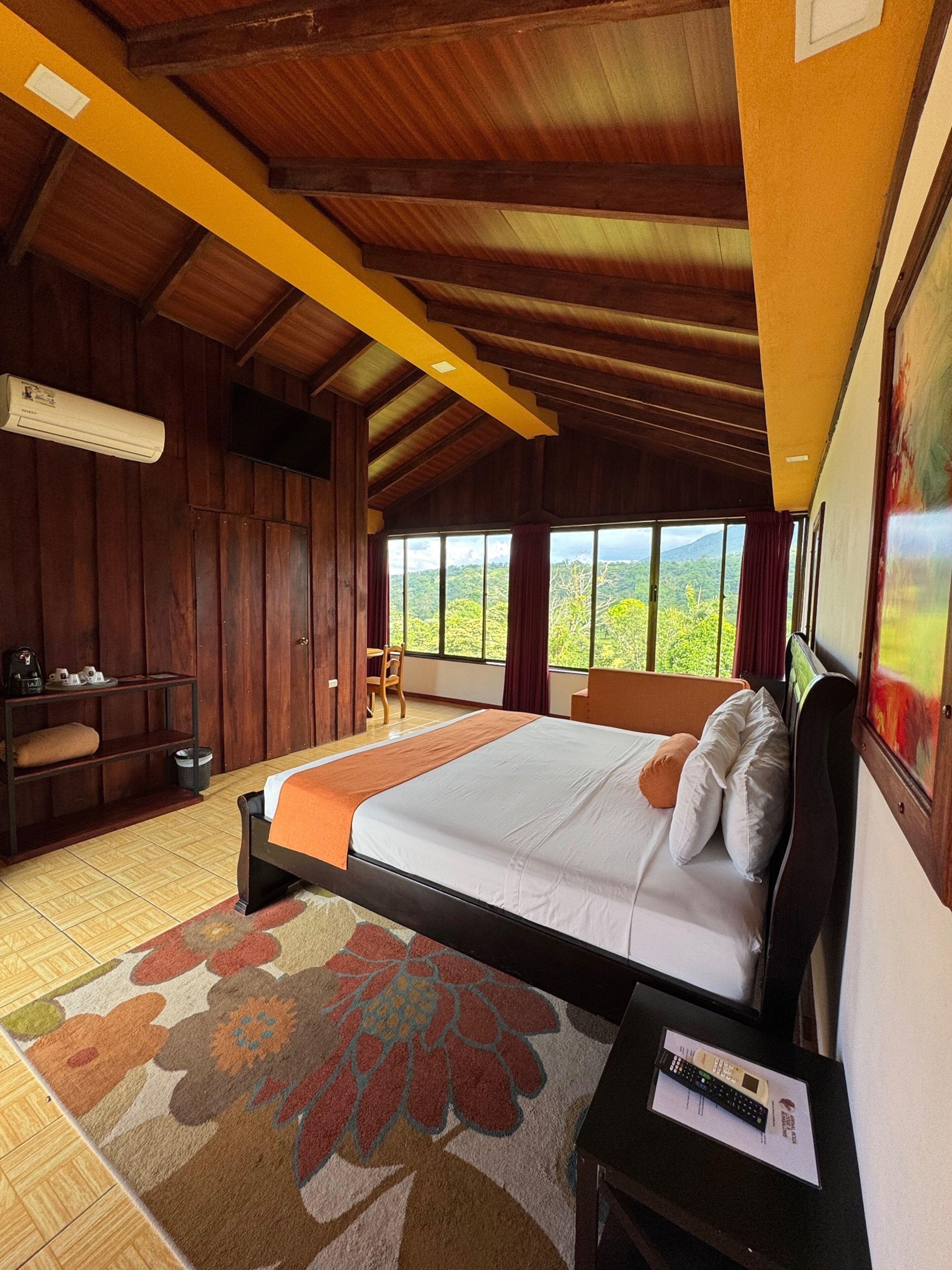 A bedroom with a large bed, wooden walls, a window with a view, and a small table with a remote control.