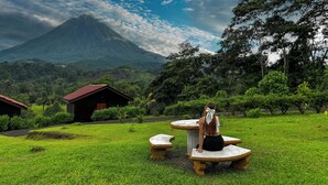 A grassy area with two stone benches and a person sitting on one. In the background, there is a mountain and a few buildings.