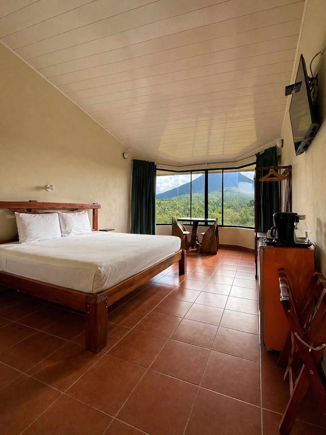 A bedroom with a bed, a table, chairs, a TV, and a view of a mountain.