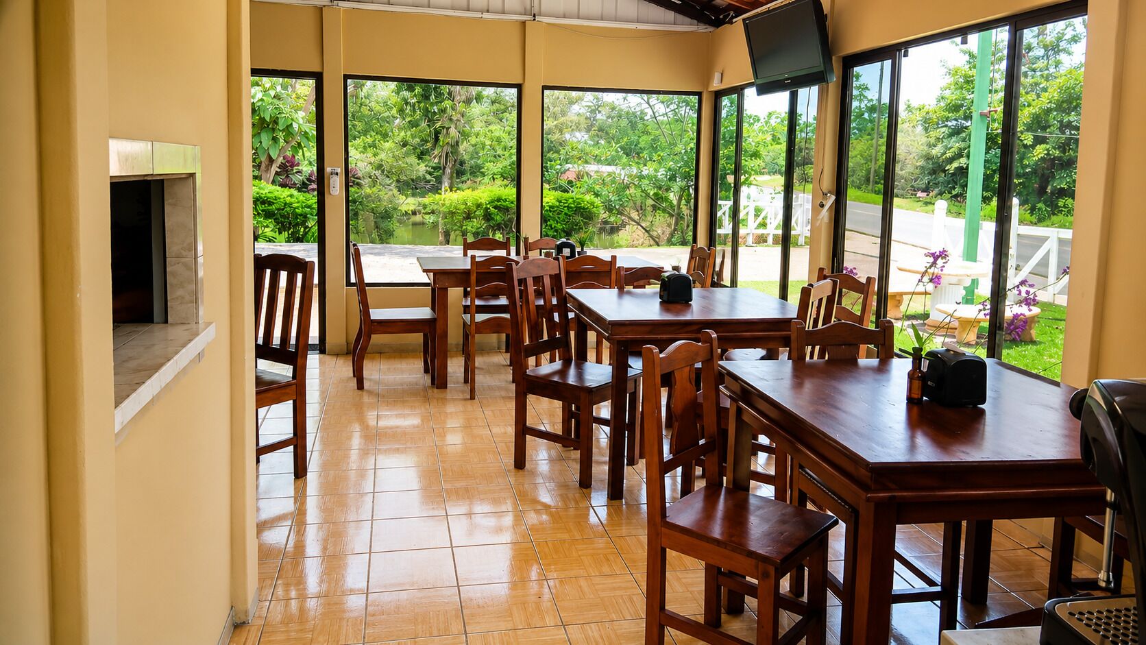 A dining area with wooden tables and chairs, large windows with a view of greenery, and a flat-screen TV mounted on the wall.