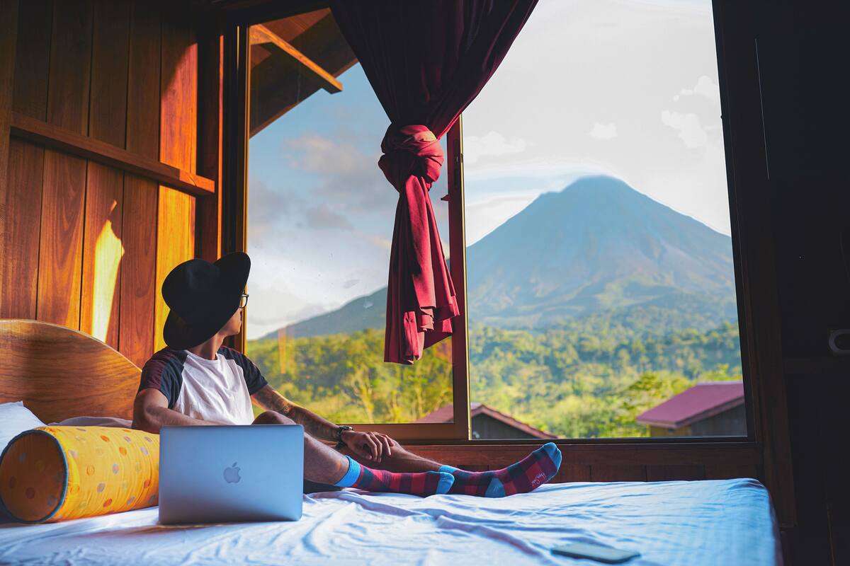 Una persona seduta su un letto con un portatile, una finestra che si affaccia su montagne e una tenda rossa.
