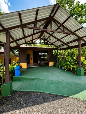 A covered reception area with a bench, a trash bin, and potted plants.