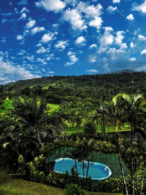 A lush landscape with a swimming pool surrounded by tropical vegetation and a distant mountain range under a partly cloudy sky. - Arenal Roca Lodge (La Fortuna)
