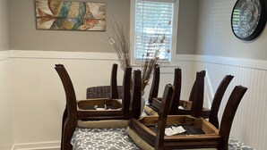 A dining area with a wooden table and chairs, a patterned tablecloth, a wall clock, and a window with blinds.