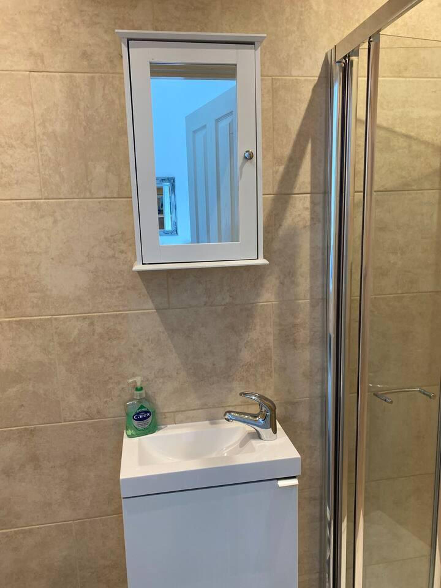 Ein Badezimmer mit einem weißen Schrank über einem Waschbecken, einer Duschkabine aus Glas und einer Flasche Carex Handseife.