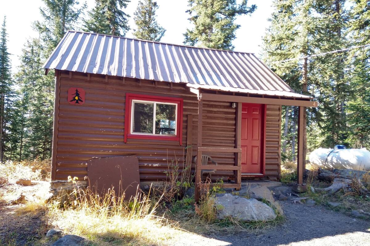 A small wooden cabin with a red door and window, surrounded by trees and rocks.