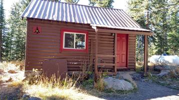 Une petite cabane en bois, avec une porte et une fenêtre rouges, entourée d’arbres et de rochers.