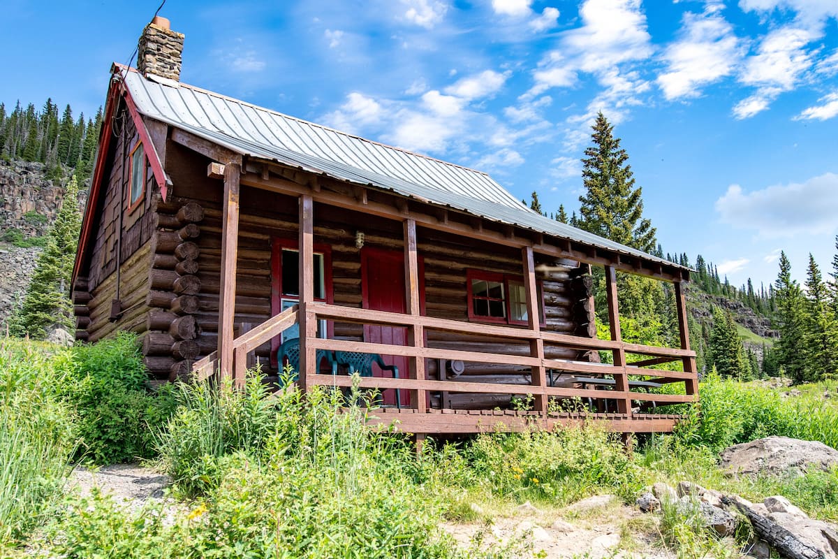 A rustic log cabin with a metal roof, wooden porch, and a scenic mountainous backdrop.