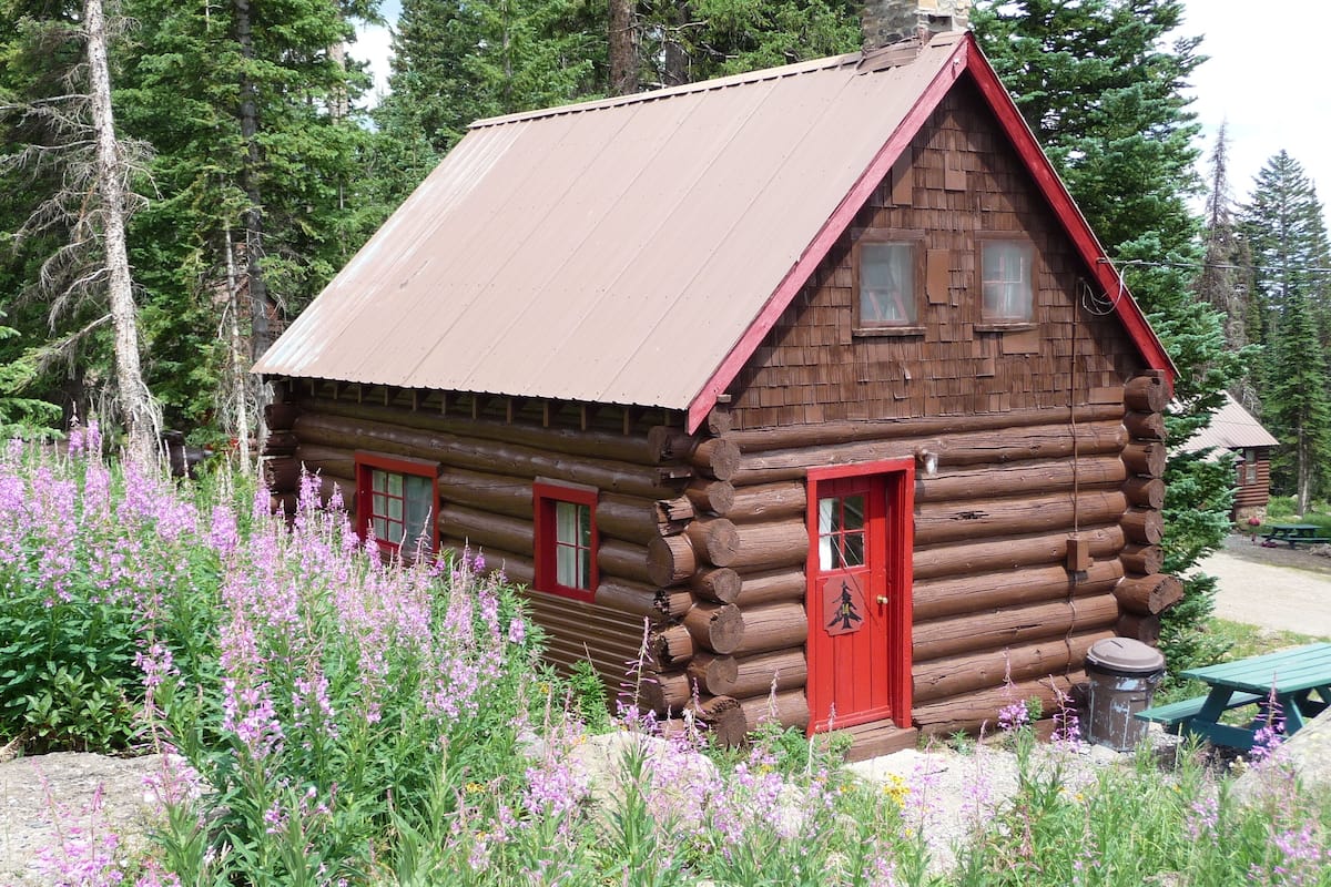 A log cabin with a red door and windows, surrounded by wildflowers and trees.
