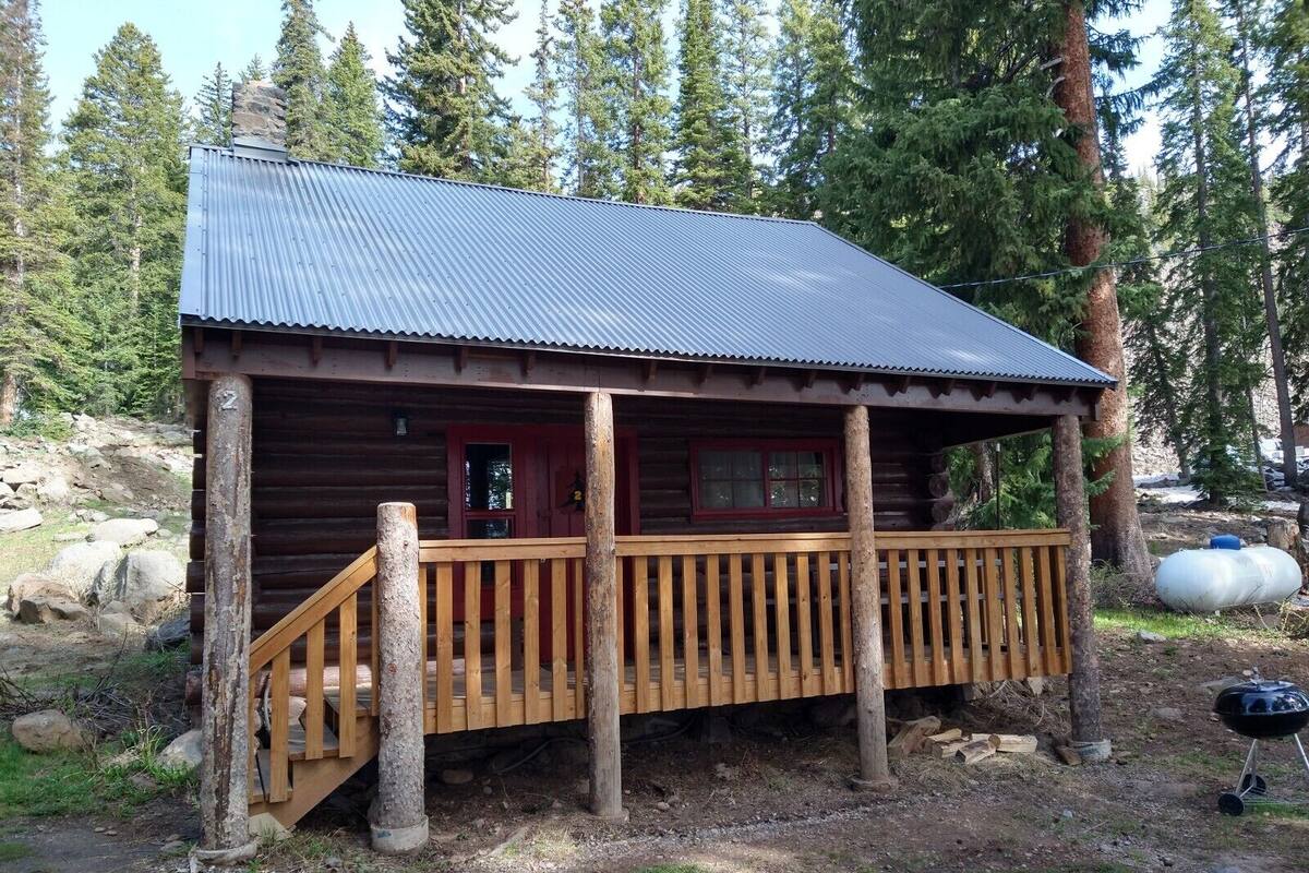 A rustic cabin with a wooden deck and metal roof, surrounded by trees.