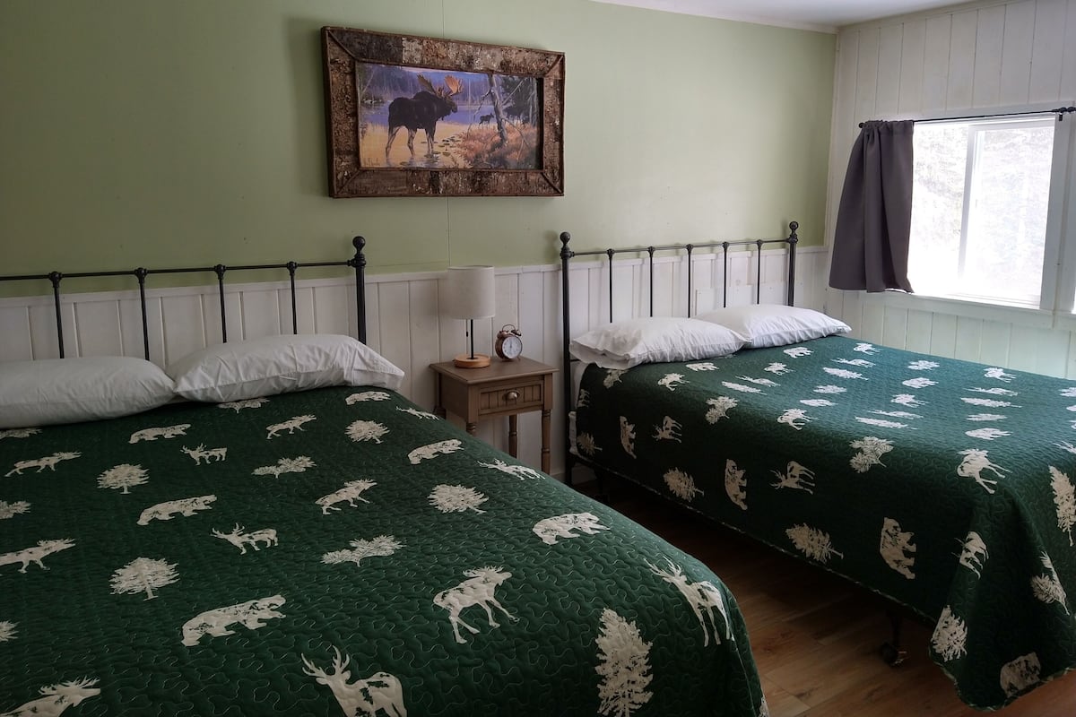 Two beds with green bedding, a nightstand with a lamp and clock, a painting on the wall, and a window with curtains.