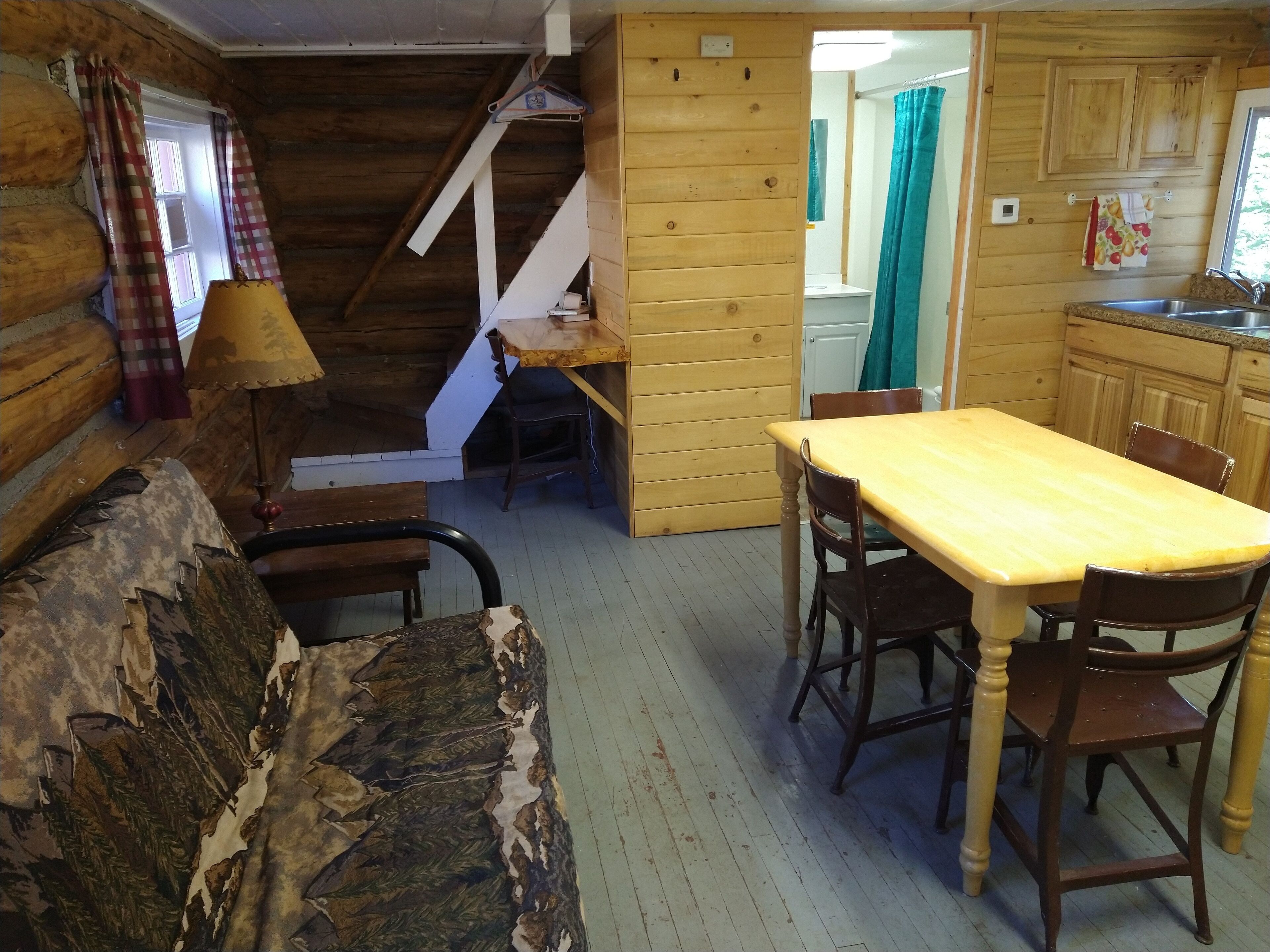 A rustic cabin interior with a wooden dining table, chairs, a sofa, and a kitchen area with wooden cabinets and a sink.