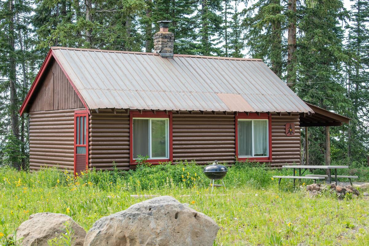 A rustic log cabin with a metal roof, two windows with curtains, a chimney, and a barbecue grill in front.