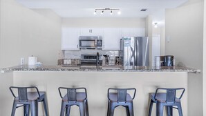Une cuisine moderne avec des armoires blanches, des appareils en acier inoxydable et un comptoir en granit. On y trouve quatre tabourets de bar en métal.