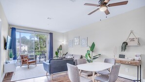 Un salon moderne avec un canapé, une table à manger et des chaises. On y trouve un ventilateur de plafond, un téléviseur et une grande fenêtre avec des rideaux.