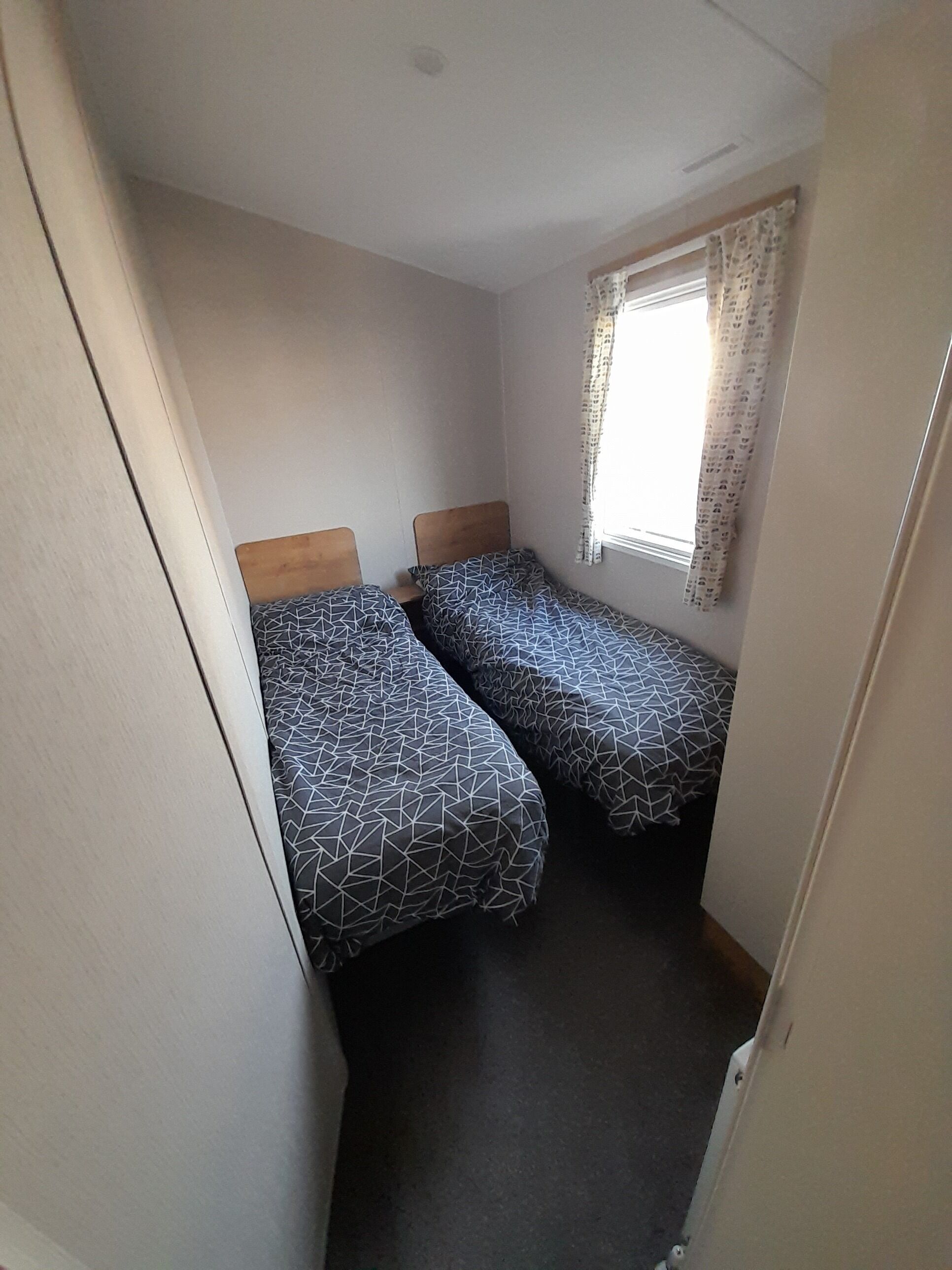 A small, narrow room with two single beds, a window with patterned curtains, and a plain white wall.