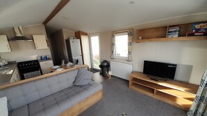 Ett kompakt vardagsrum med ett litet kök, en grå soffa, en tv och ett fönster med mönstrade gardiner.