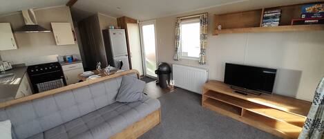 A compact living space with a kitchenette, a grey sofa, a television, and a window with patterned curtains.