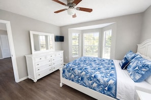 A bedroom with a bed, a dresser, a ceiling fan, and large windows.