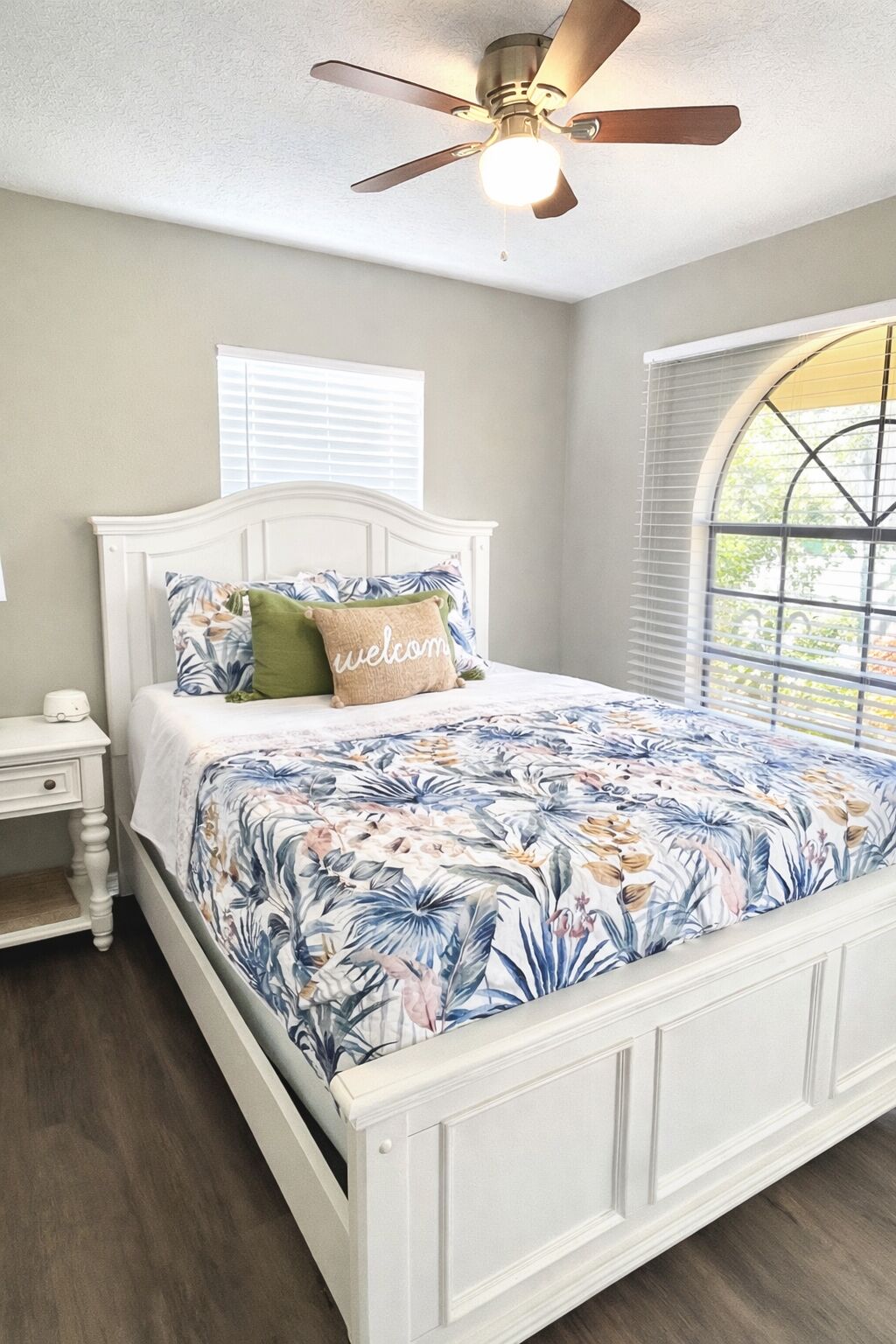 Un dormitorio con cama blanca, ropa de cama floral, ventilador de techo y ventana con persianas.