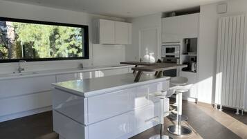 Une cuisine moderne avec des meubles blancs, un grand îlot central doté de tabourets de bar et une fenêtre donnant sur un espace verdoyant.