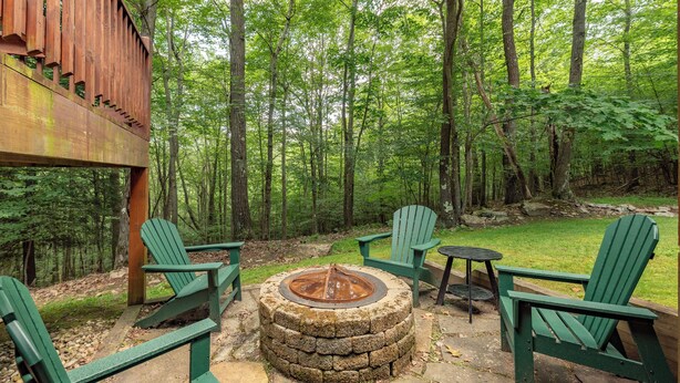 Un foyer circulaire en pierre, entouré de fauteuils Adirondack verts, se trouve dans un espace boisé agrémenté d’une pergola en bois.