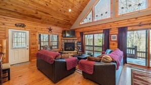 L’intérieur d’une cabane en rondins, avec une cheminée, deux canapés, un téléviseur et de grandes fenêtres.