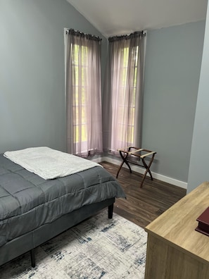 A bedroom with a bed, a chair, a wooden desk, and sheer curtains.