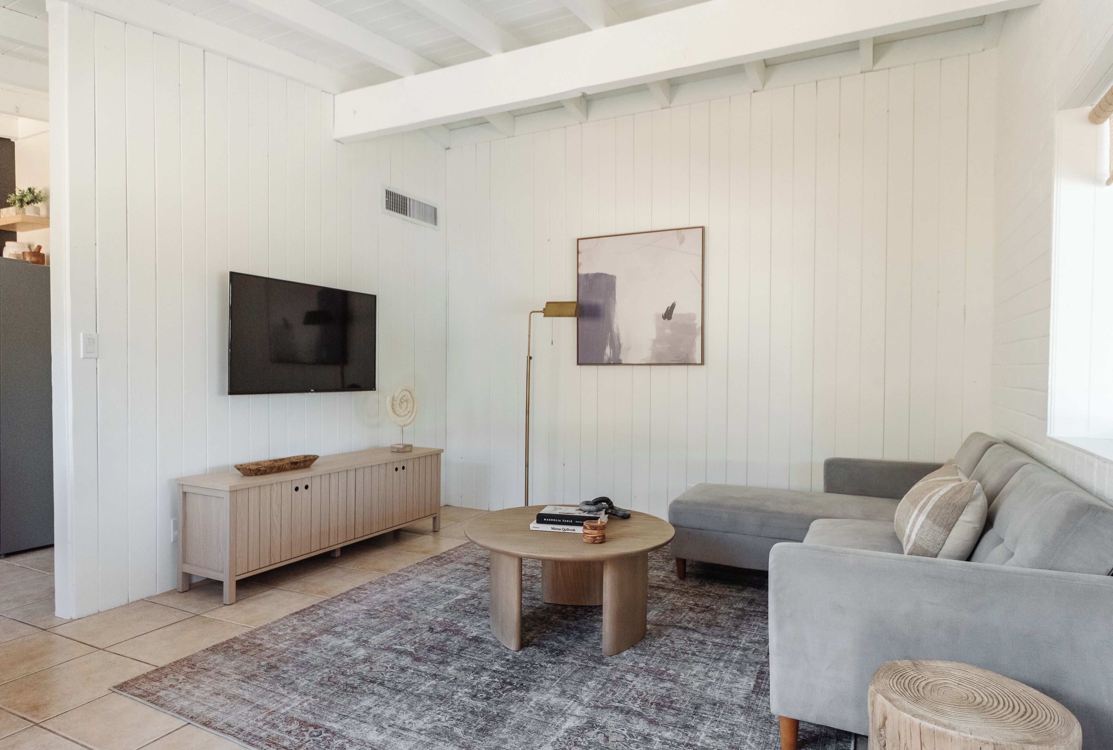 Ein modernes Wohnzimmer mit einem grauen Sofa, einem runden Couchtisch, einer Holztruhe, einem Flachbildfernseher und einem gerahmten Kunstwerk an der Wand.