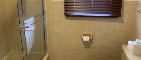 A bathroom with a glass-enclosed shower, a toilet, and a window with blinds.