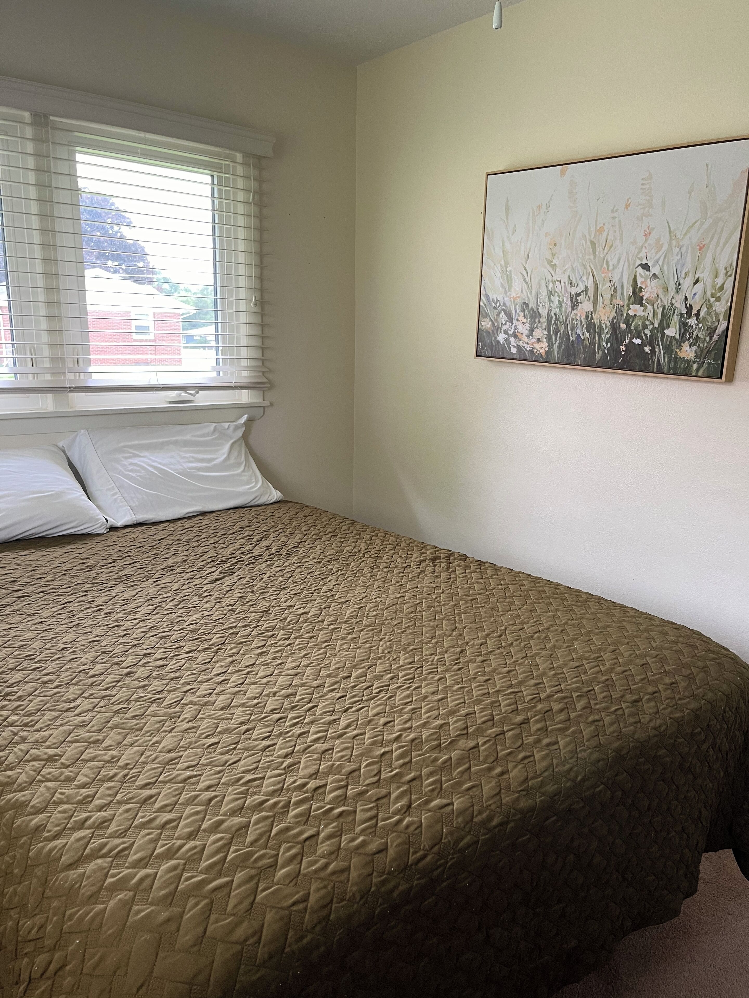 A bedroom with a bed, white pillows, a window with blinds, and a painting on the wall.