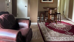 A living room with a brown leather couch, a dining table with four wooden stools, a patterned rug, and a framed painting on the wall.