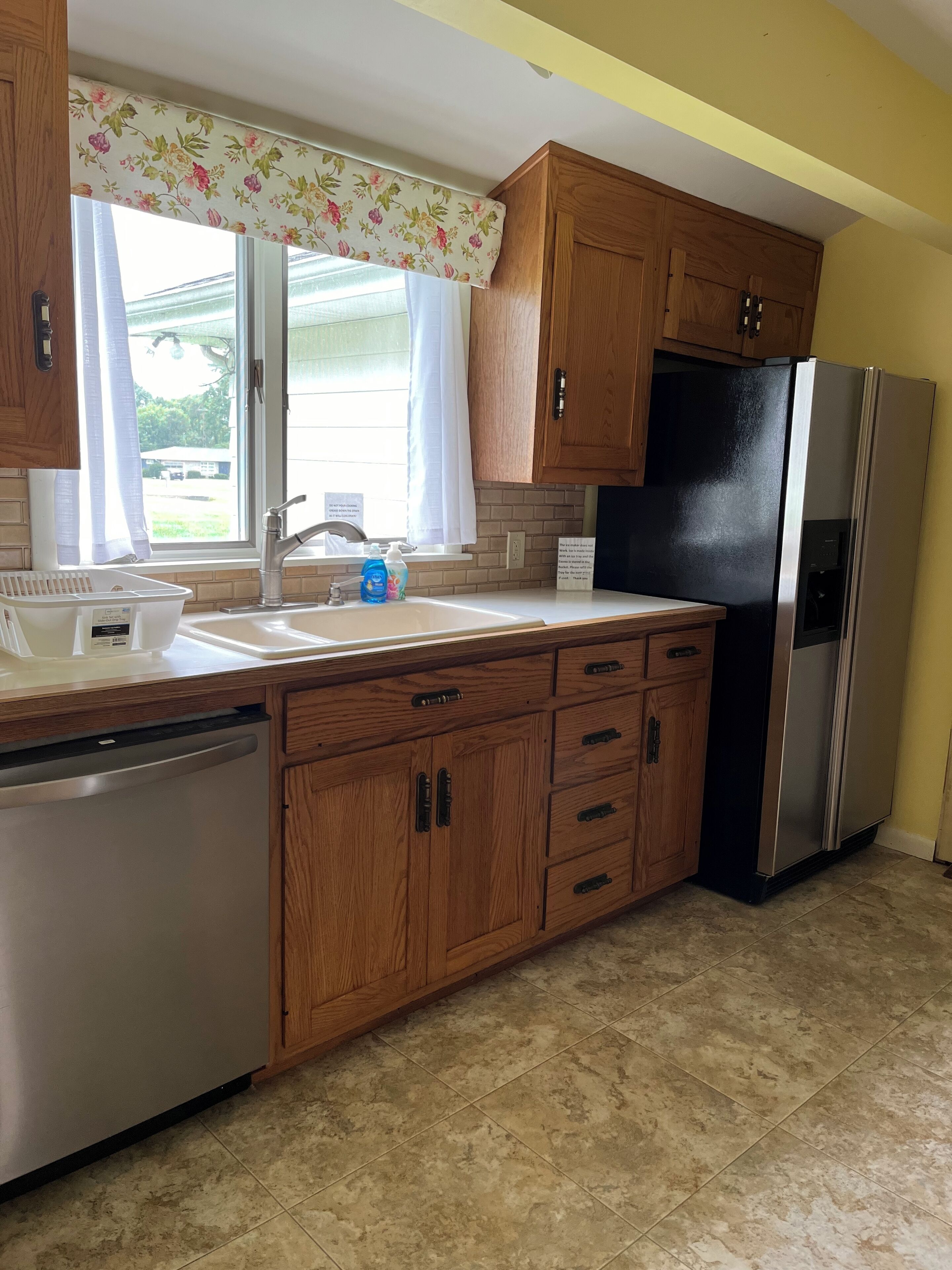 A kitchen with wooden cabinets, a stainless steel refrigerator, a dishwasher, and a window with a floral curtain.