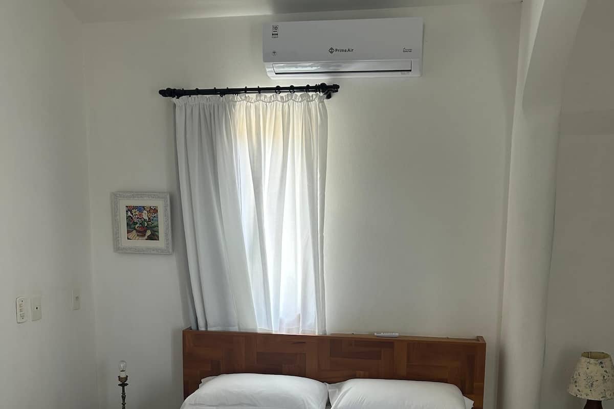 Ein ordentlich bezogenes Bett mit weißen Leinentüchern, ein Nachttisch mit Lampe, eine Wand-Klimaanlage und ein Fenster mit durchsichtigen Vorhängen.