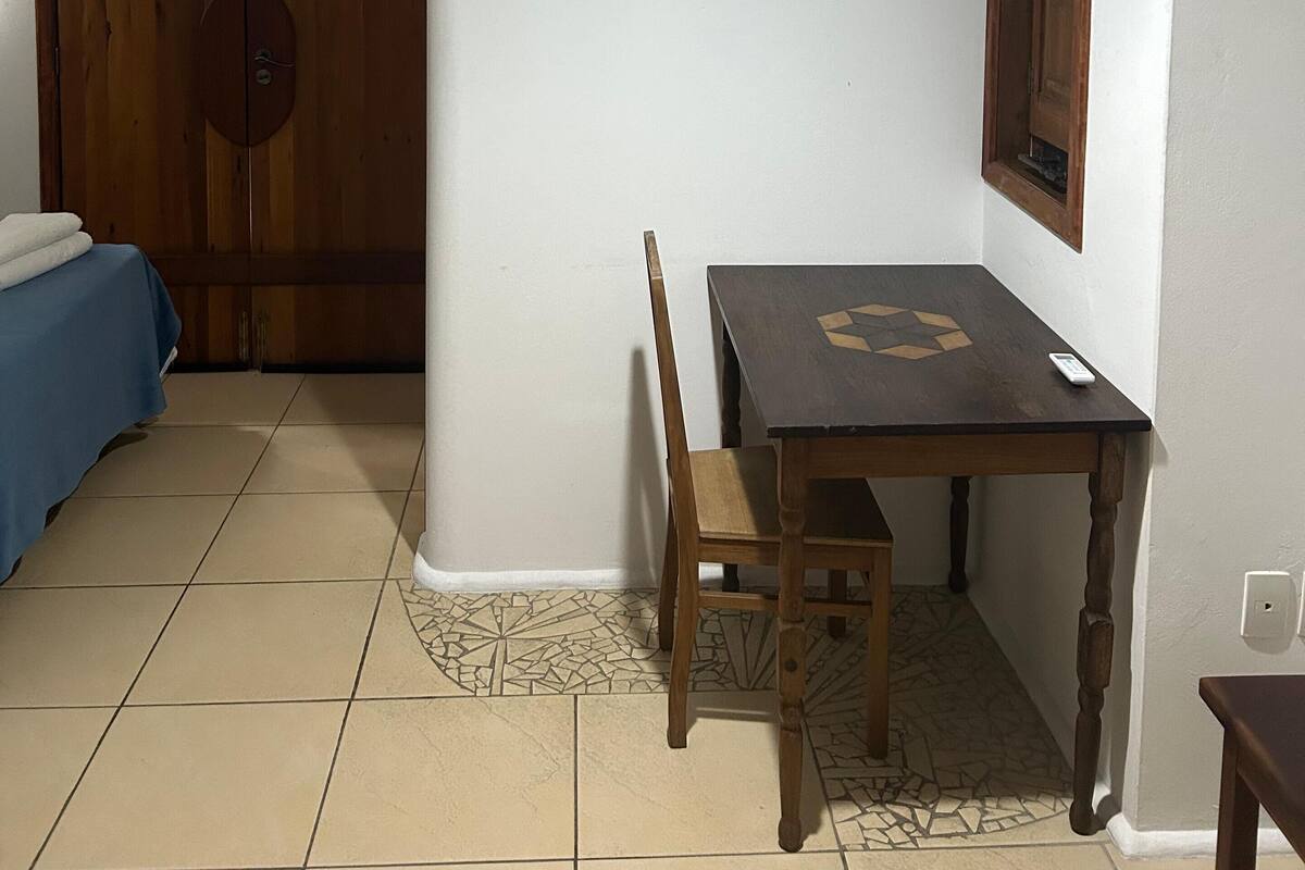 Ein Zimmer mit Holztür, einem Bett mit blauer Bettdecke, einem Tisch mit Stuhl und einer Klimaanlage an der Wand.