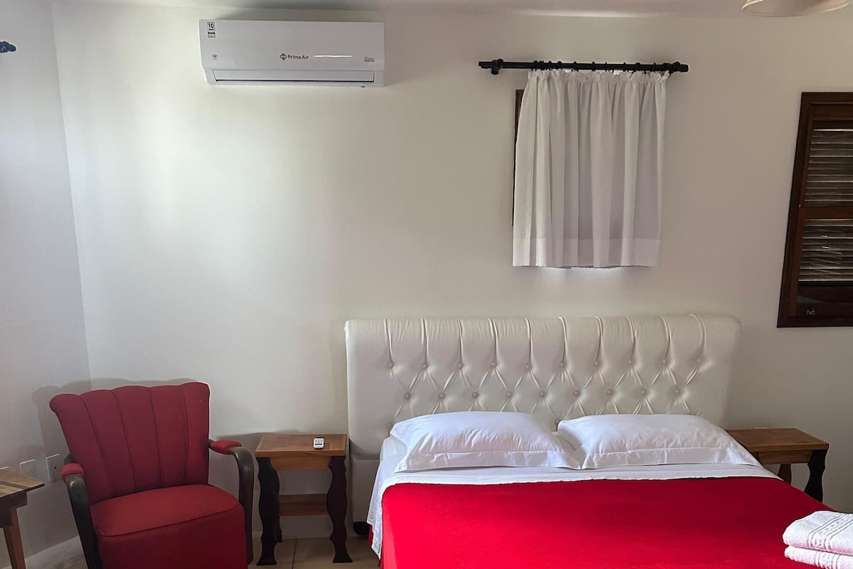 Ein Schlafzimmer mit einem Bett, einem roten Sessel, einem Nachttisch aus Holz, einem kleinen Tisch und einer Wand-Klimaanlage.