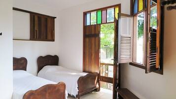 Una habitación con dos camas de madera, un banco, una ventana con contraventanas y un estante en la pared.