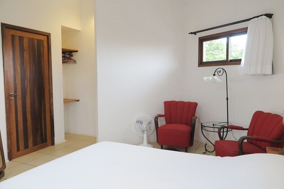 A room with a bed, two red chairs, a small table, a standing fan, a window with curtains, and a wooden door.