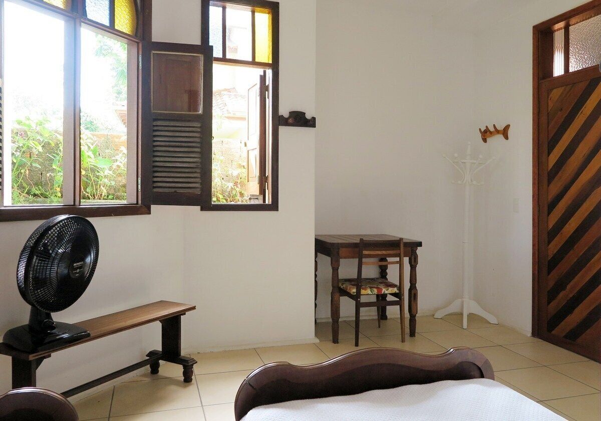A room with a bed, a desk, a chair, a fan, and a window with a view of greenery.